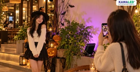 Hai bạn trẻ chụp ảnh tại góc check-in Halloween có ánh sáng màu trang trí và đèn vàng ấm.