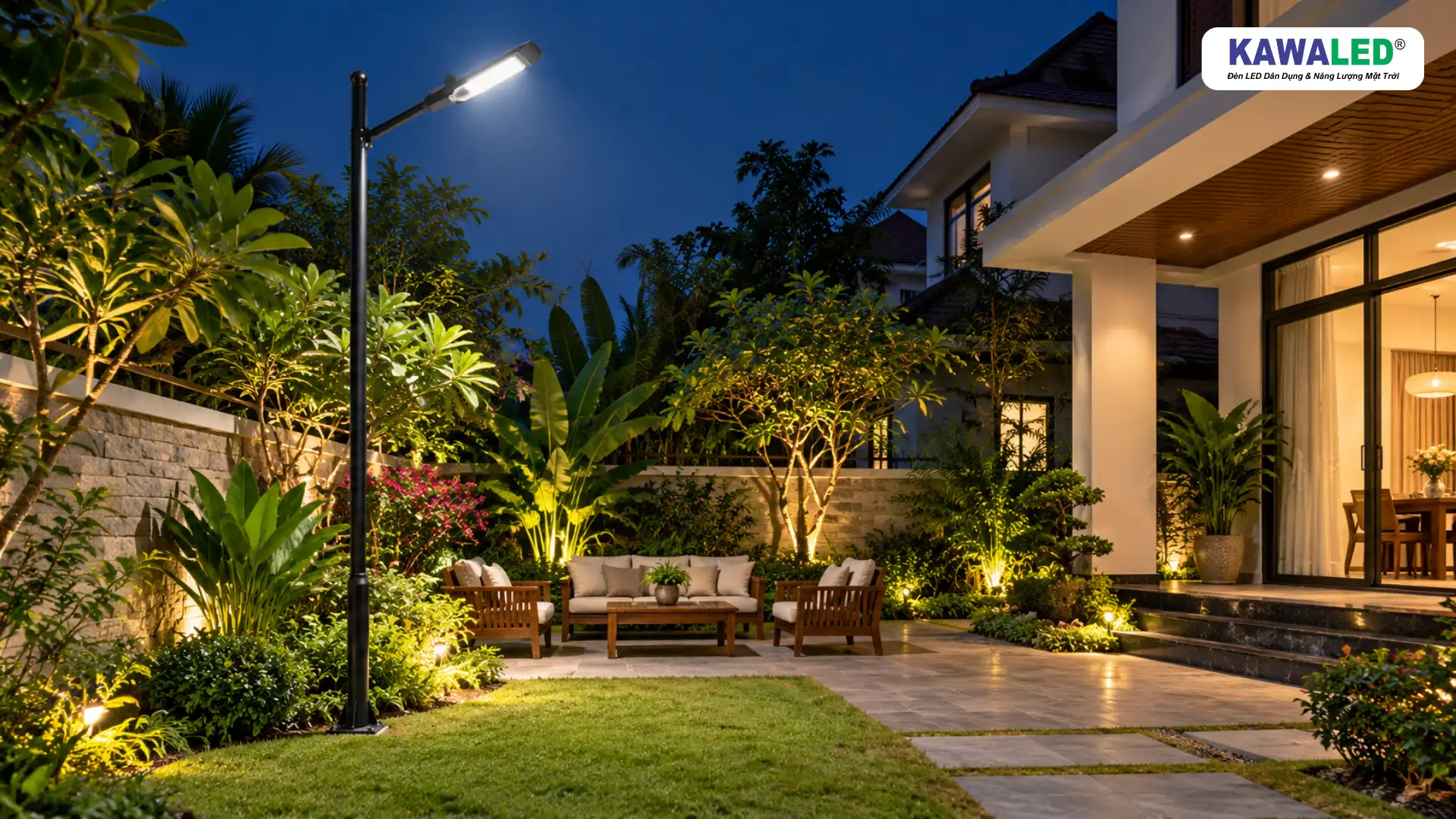Đèn chiếu sáng sân vườn trên trụ đen cạnh thảm cỏ, cây xanh và bộ bàn ghế thư giãn ngoài trời.