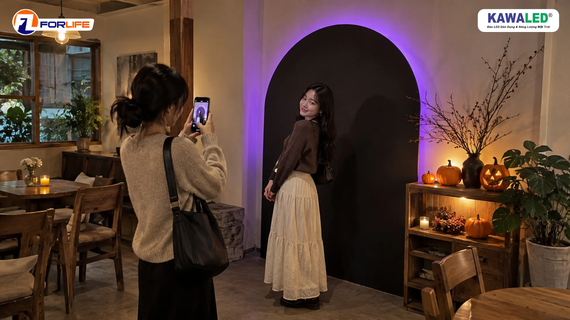 Hai khách nữ chụp ảnh tại góc check-in Halloween với phông vòm hắt tím và kệ bí ngô ánh vàng.