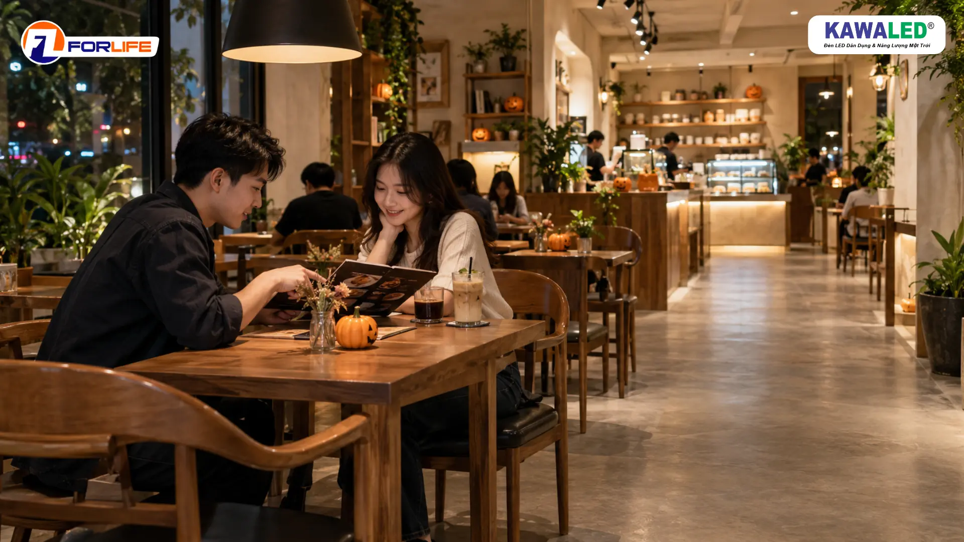 Hai khách xem menu tại bàn gỗ dưới đèn thả, bên cạnh là lối đi rộng trong quán cà phê Halloween.